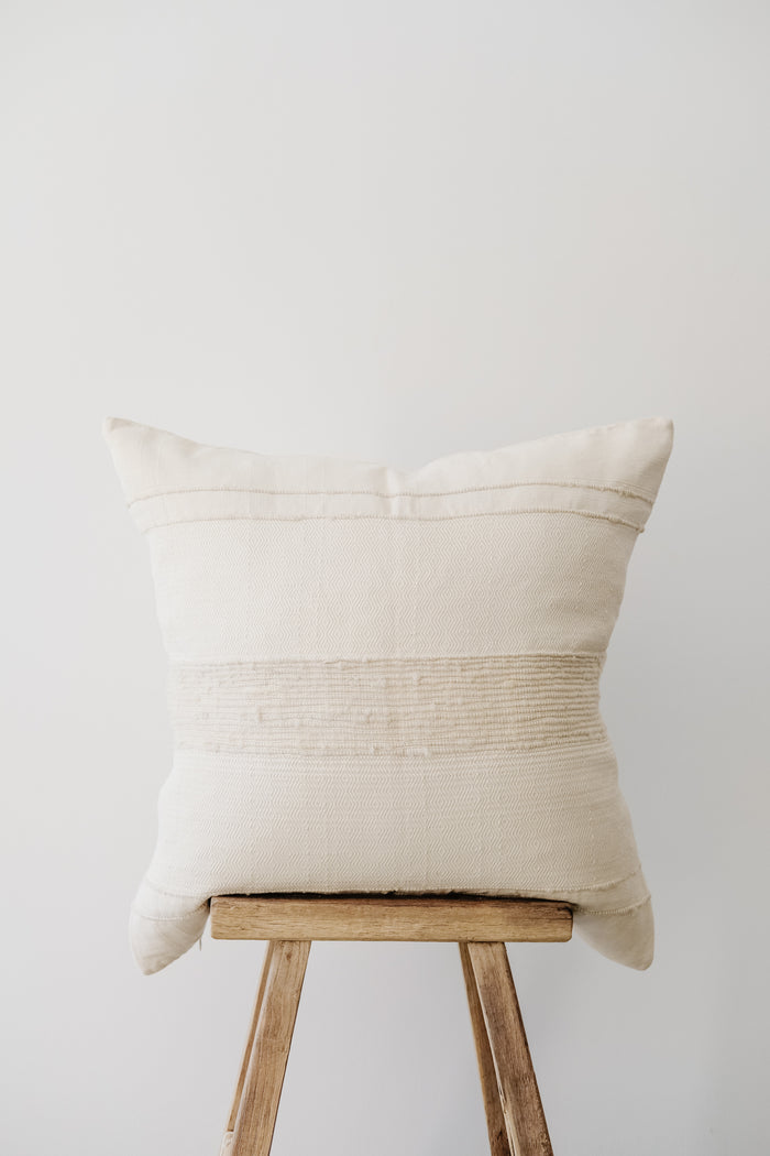 Front view of No. 50 Azulina Ivory with Ivory Stripe Handwoven Columbian Pillow on an Antique Stool against a white wall. - Saffron + Poe