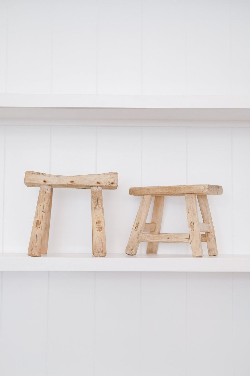 Two petite antique elmwood stools with mortise and tenon joinery on a white shelf