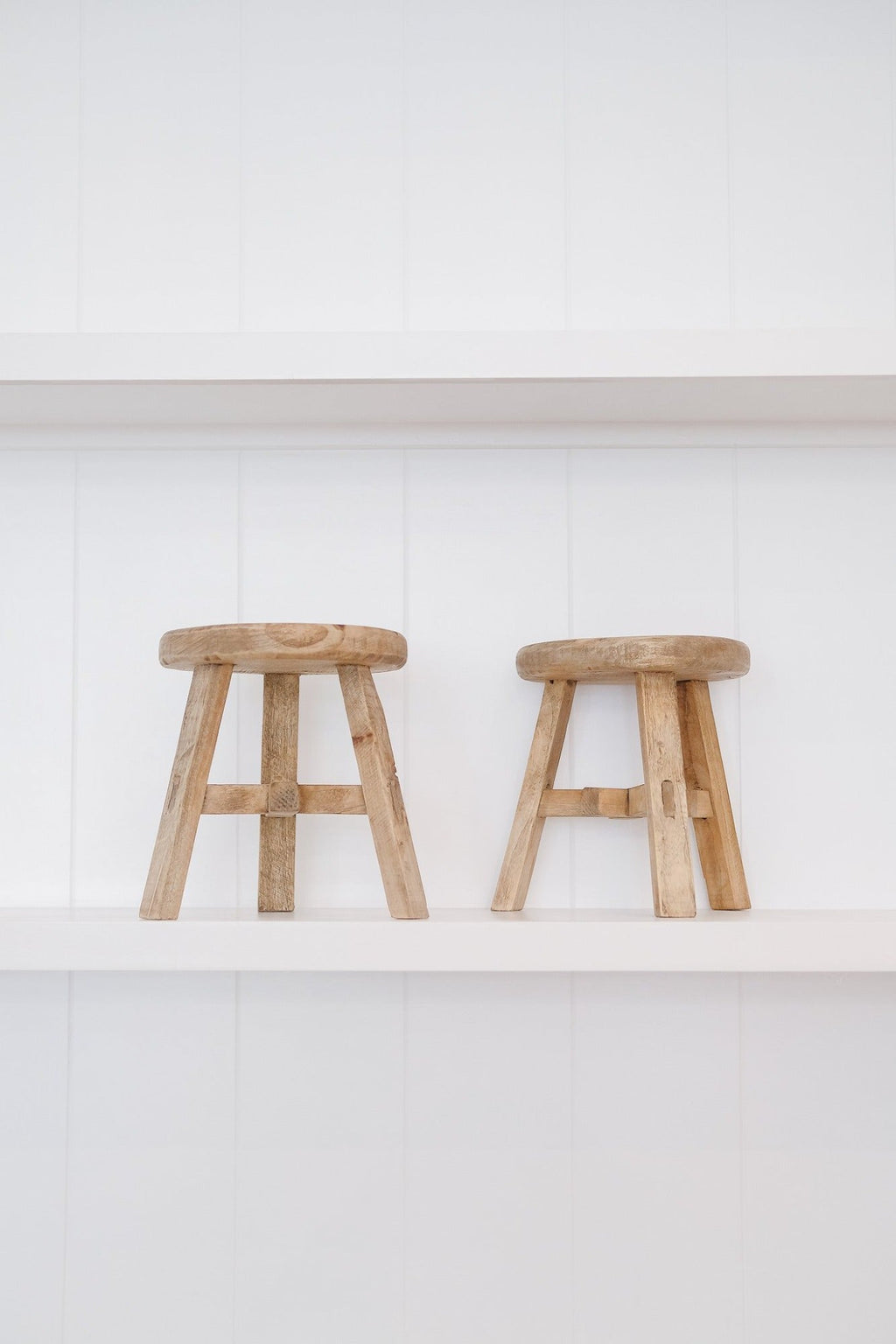 Two petite vintage wooden stools on white shelves against white panel wall