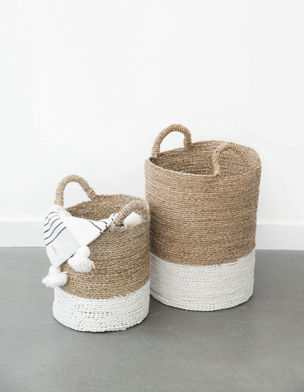 Two-toned structured hyacinth storage baskets with handles on a gray floor, one holding a white cloth with tassels