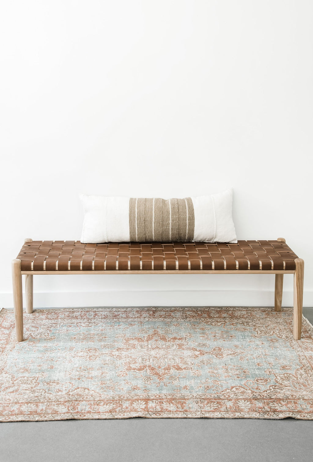 Woven leather bench with teak frame and saddle-colored leather straps on patterned rug with striped pillow