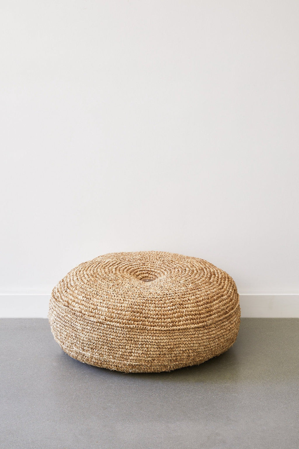 Natural woven pouf made from handwoven water hyacinth fibers, placed on a gray floor against a white wall.