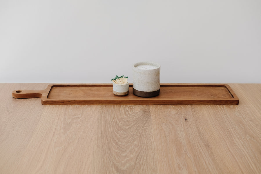 Styled Hand Carved Teak Serving Tray with an Uzumati Ceramic Candle and a Hand Thrown Match Striker on an Oak Dining Table against a white wall. - Saffron and Poe