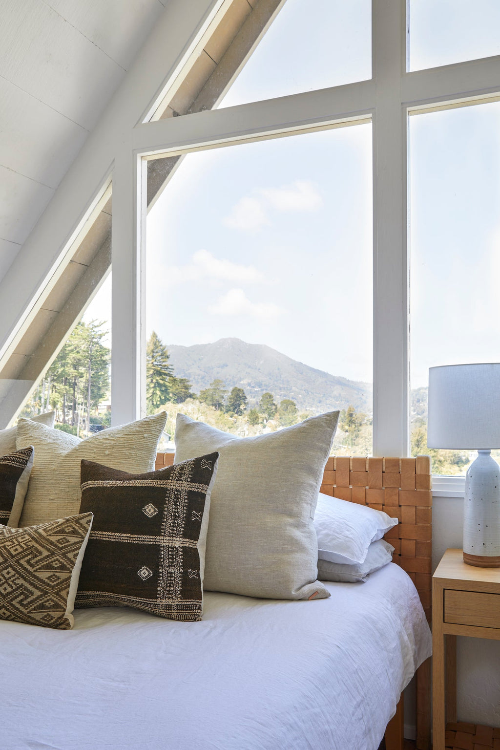 Bright bedroom with white linen duvet set and decorative pillows by large window with mountain view