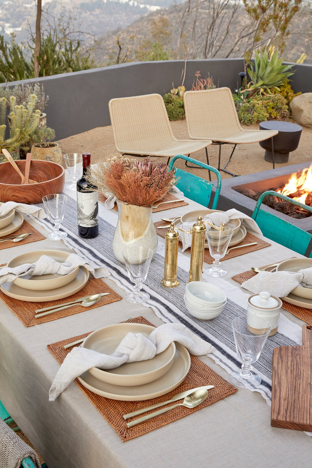 Outdoor dining table set with natural linen napkins, beige plates, gold utensils, woven placemats, and a fire pit in the background