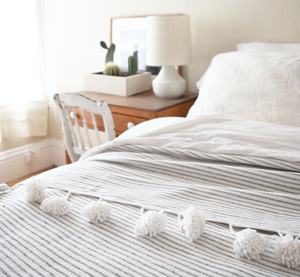 Moroccan Pom Bed Throw light grey and white striped with oversized pom tassels on a bed