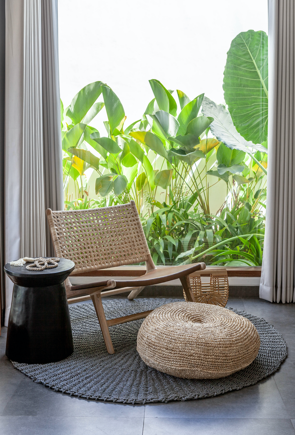 Natural woven hyacinth pouf on gray rug beside wooden chair and black side table in bright room with green plants
