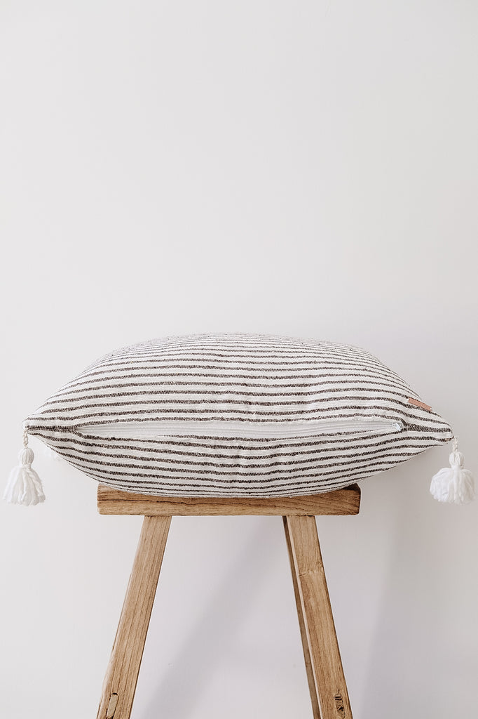 Side view of No. 40 - Moroccan Pom Pom Striped Pillow - Dark Grey on a Chinese Stool against a white wall. - Saffron and Poe