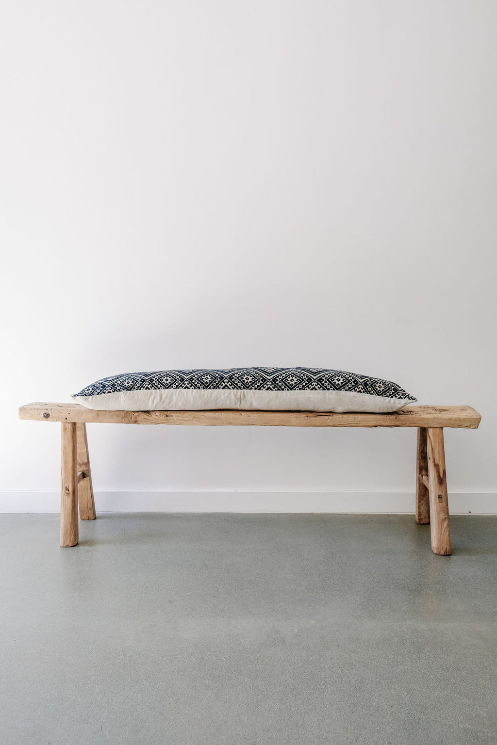 Antique Miao lumbar pillow with intricate blue and white woven patterns on rustic wooden bench against white wall