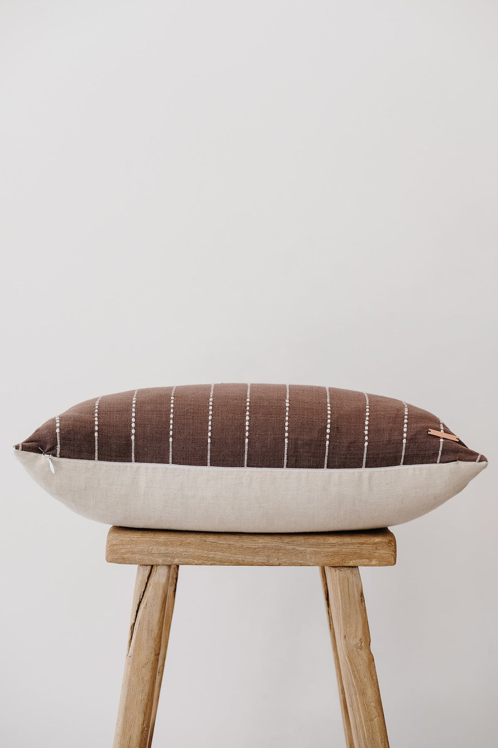 Brown embroidered lumbar pillow with linen backing on wooden stool against neutral background
