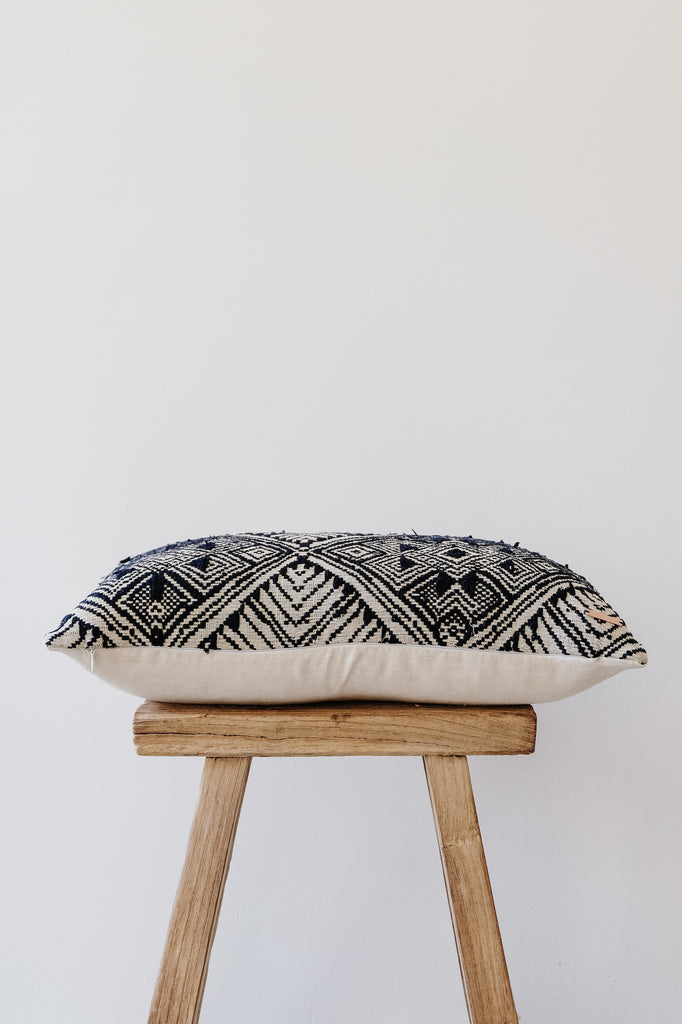 Side view of No. 21 - Antique Miao Lumbar Pillow on an Antique Stool against a white wall. - Saffron and Poe