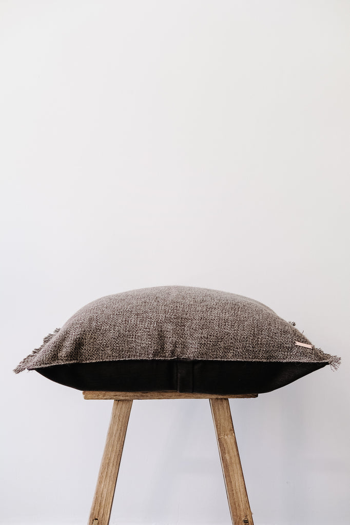 Side view of No. 29 - Baby Alpaca Pillow - Dark Brown on an Antique Stool against a white wall. - Saffron and Poe