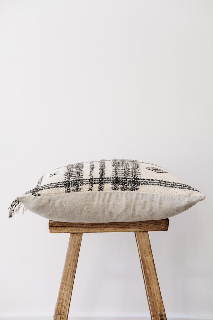 Side view of No. 30 - Handwoven Bhujodi Pillow with Fringe - Natural on an Antique Stool against a white wall. - Saffron and Poe