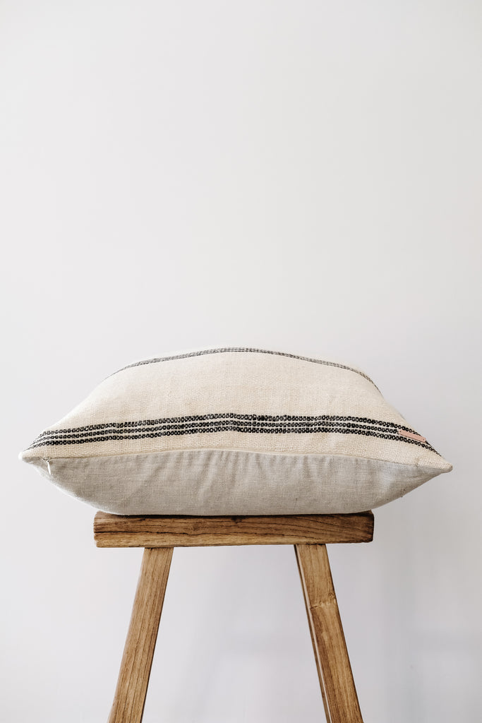 Side view of No. 31 - Handwoven Bhujodi Pillow - Natural on an Antique Stool against a white wall. - Saffron and Poe
