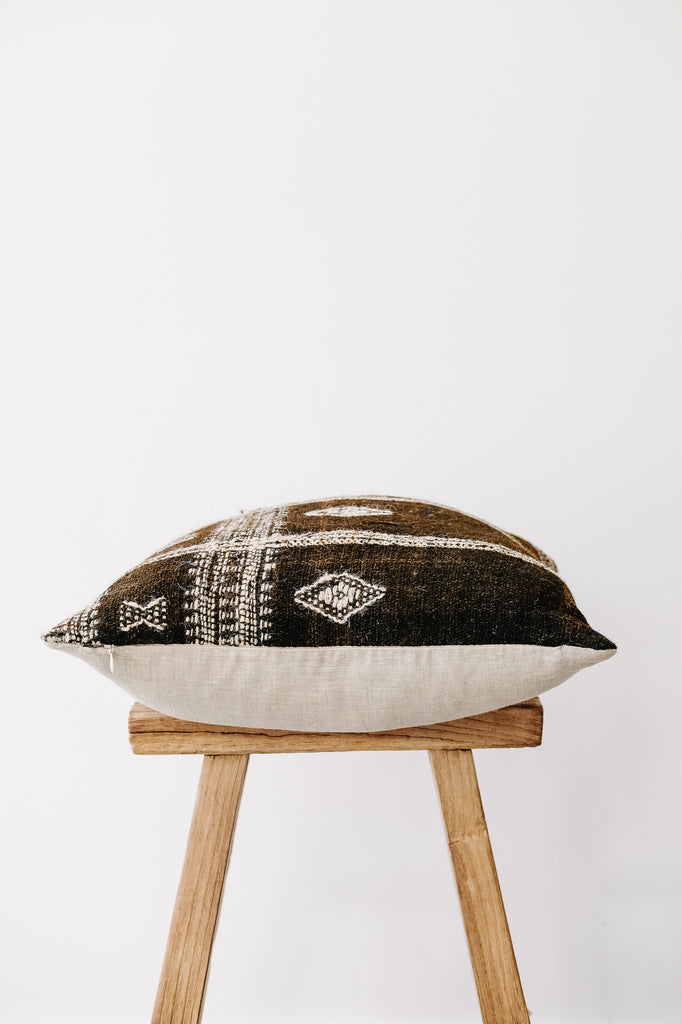 Side view of No. 33 - Handwoven Bhujodi Pillow - Dark Brown on an Antique Stool against a white wall. - Saffron and Poe