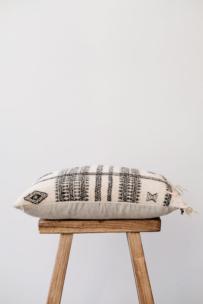 Side view of No. 36 - Handwoven Bhujodi Lumbar Pillow with Fringe - Natural on an Antique Stool against a white wall. - Saffron and Poe