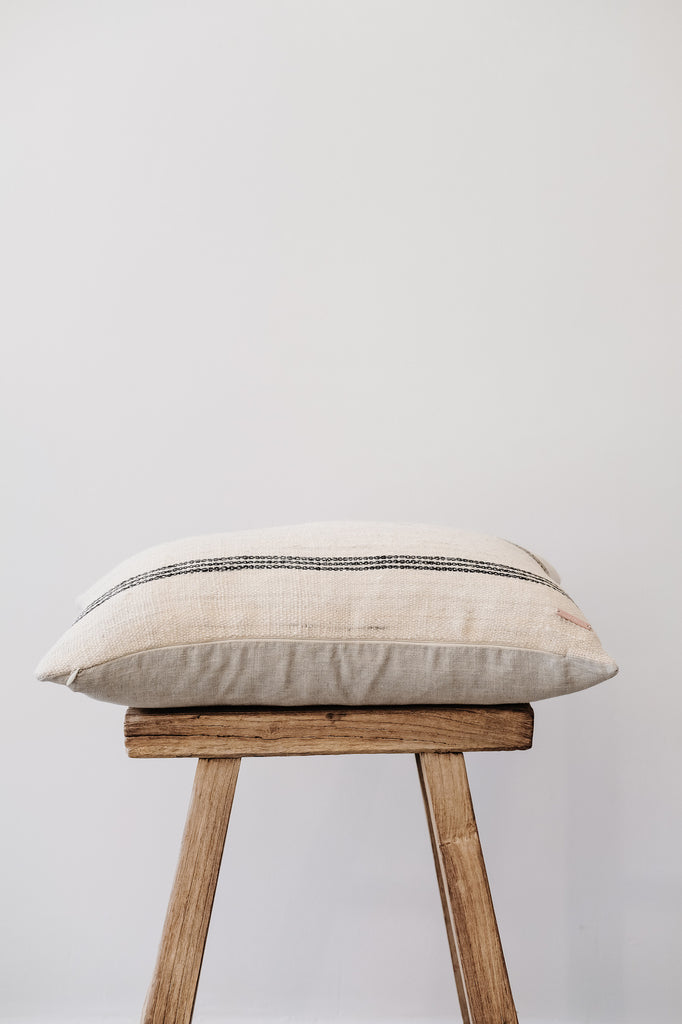 Side view of No. 37 - Handwoven Bhujodi Lumbar Pillow - Natural on an Antique Stool against a white wall. - Saffron and Poe