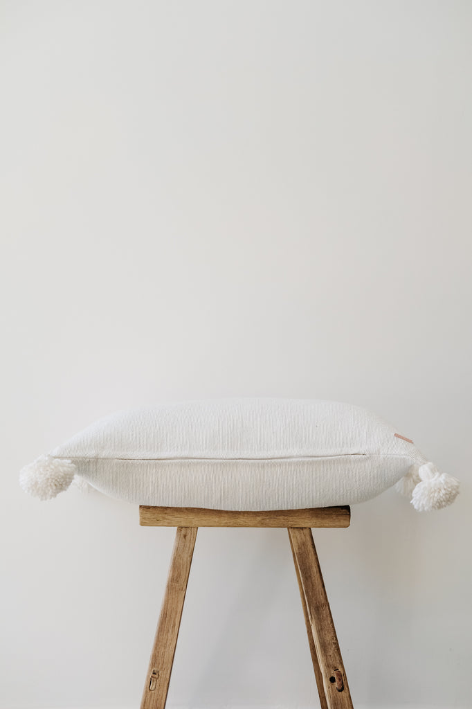 Side view of No. 38 - Moroccan Pom Pom Pillow - White on a Chinese Stool against a white wall. - Saffron and Poe