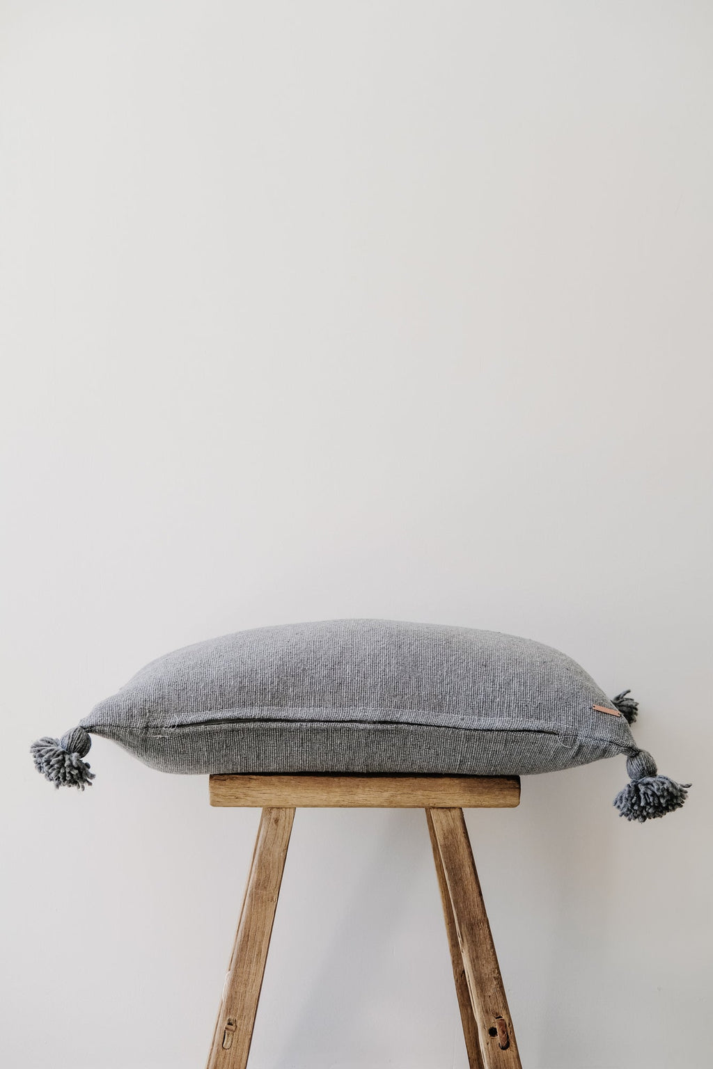 Grey Moroccan pom pillow with tassels on wooden stool against white background