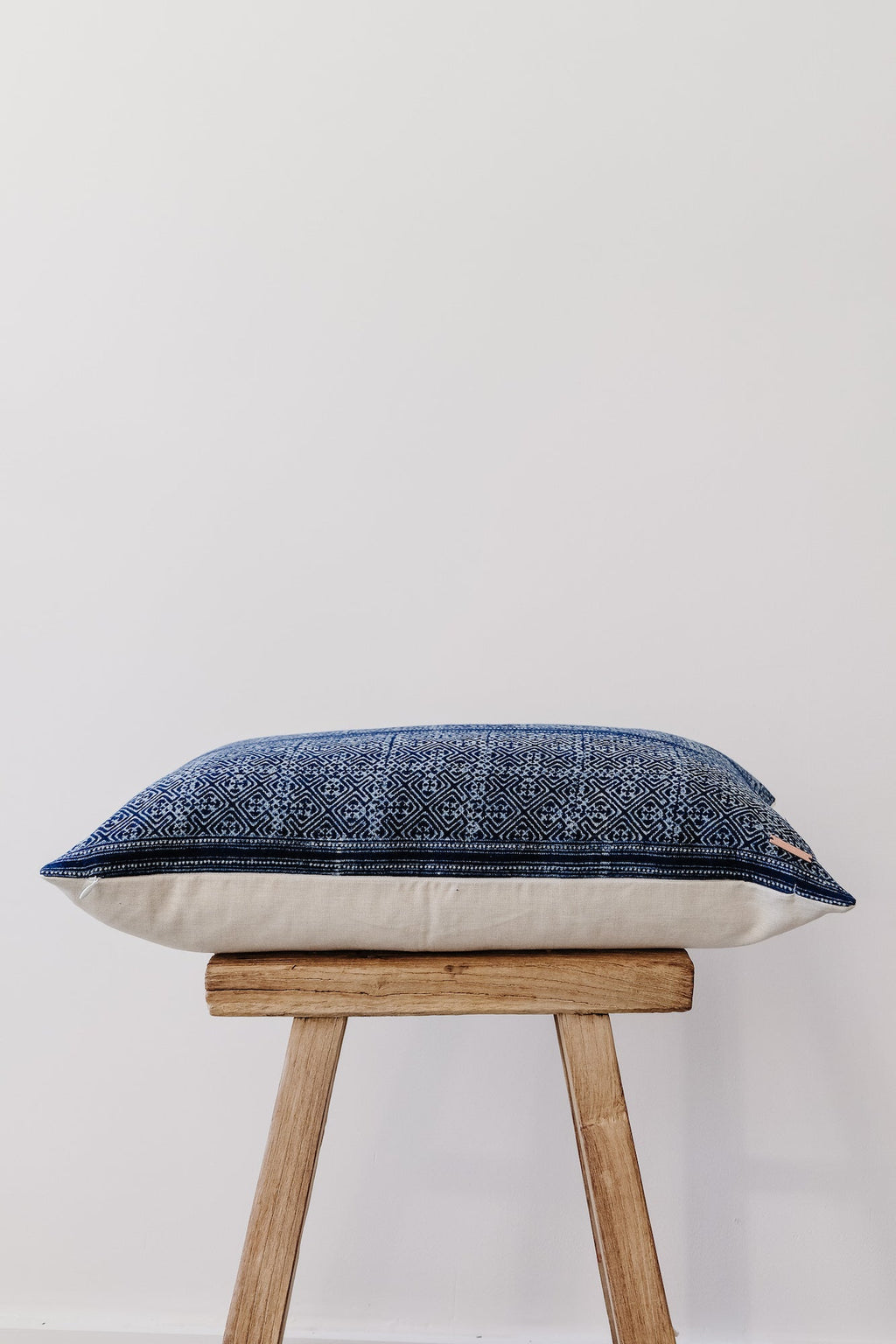 Hmong Batik lumbar pillow with indigo blue intricate patterns on wooden stool against white wall