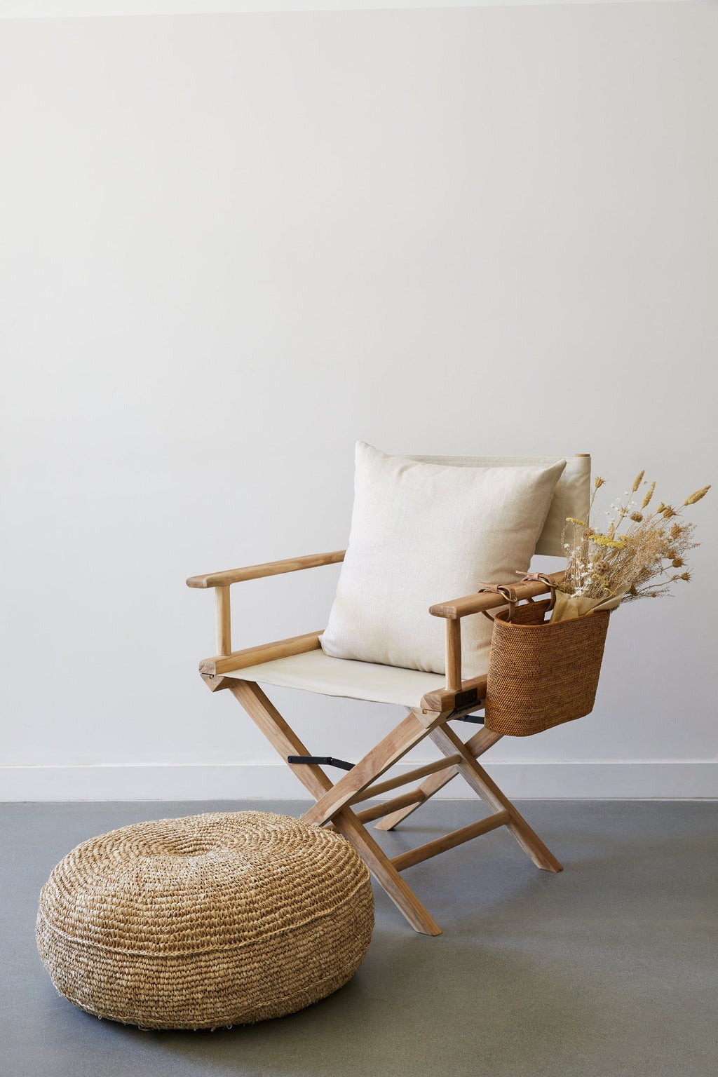 Cream linen chevron pillow on wooden director’s chair with woven basket and decorative dried flowers next to a round woven pouf against white wall