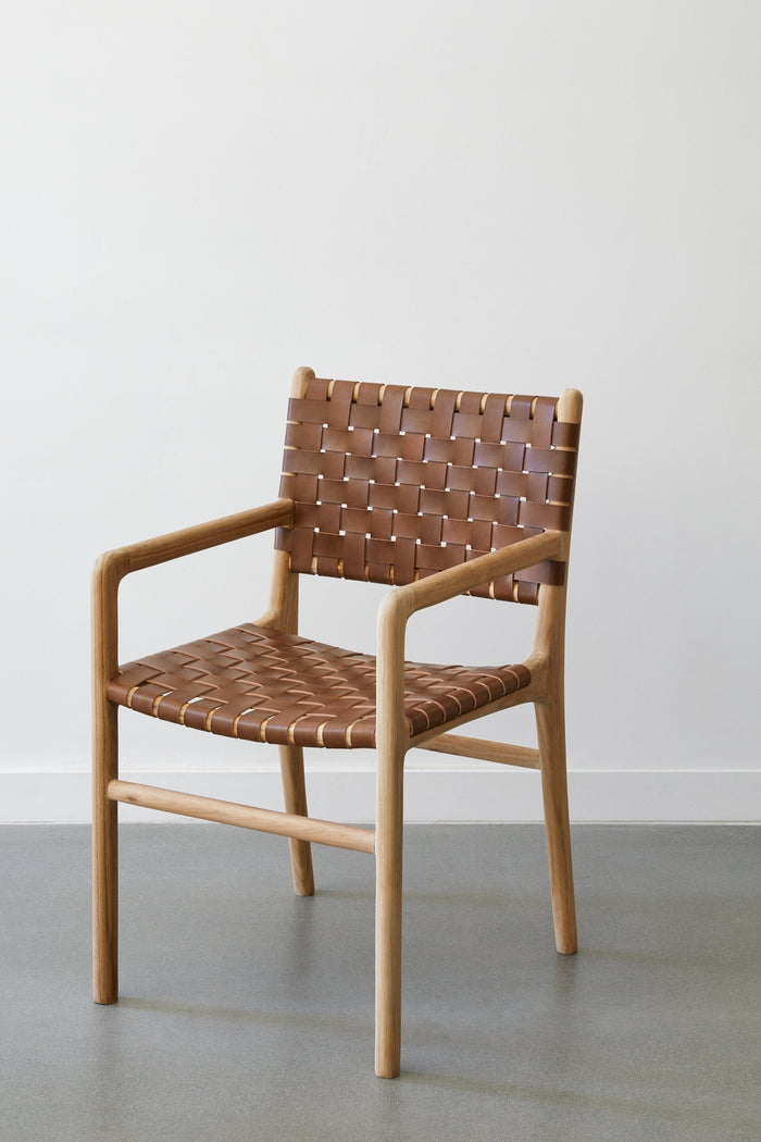 Side view of the woven leather strap dining arm chair in saddle against white background and concrete floors. - Saffron and Poe