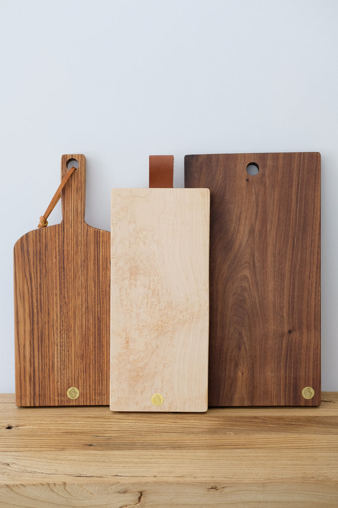 Front view of Studio Inko Zebrawood Cutting and Serving Board  stacked with Bird's Eye Maple and Walnut boards against a white wall. - Saffron and Poe