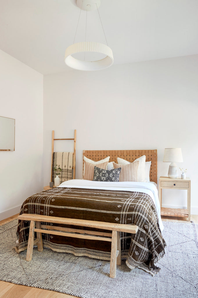 Styled Antique Miao PIllow with Beige Embroidered Pillow in a neutral tone bedroom with Handwoven Bhujodi Blanket and a Woven Leather Strap Headboard and an Antique Chinese Bench and Oak and Teak Leather Strap Side Tables. - Saffron and Poe