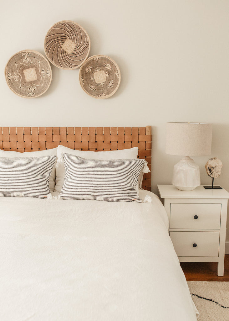 Styled Dark Grey Stripe Moroccan Pom Pillow with a Woven Leather Strap Headboard and Ilala Palm Wall Baskets in a bright bedroom. - Saffron and Poe