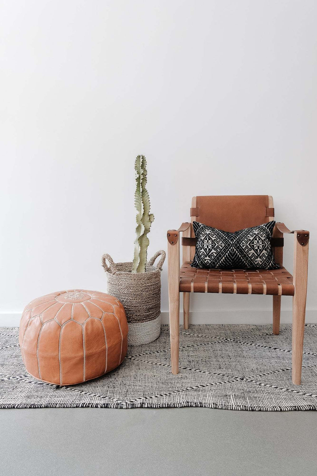 Embroidered Moroccan leather pouf next to wooden chair with black patterned pillow and woven plant basket on gray rug