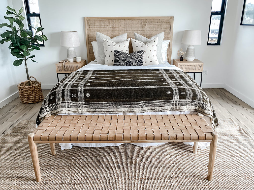 Full front view of styled bed with rattan headboard, nightstands, white walls, hardwood floors, jute area rug, woven leather strap bench in beige, indoor ficus tree in woven basket, antique Miao lumbar pillow, and Indian bhujodi bed throw in dark brown.