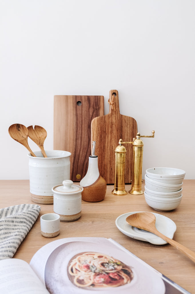 Styled Hand Thrown Ceramic Utensil Jar with Teak Tongs and the Hanselmann Pottery Collection with Studio Inko Cutting Boards and Uzumati Ceramic Olive Oil Bottle on an Oak Dining Table against a white wall. - Saffron and Poe