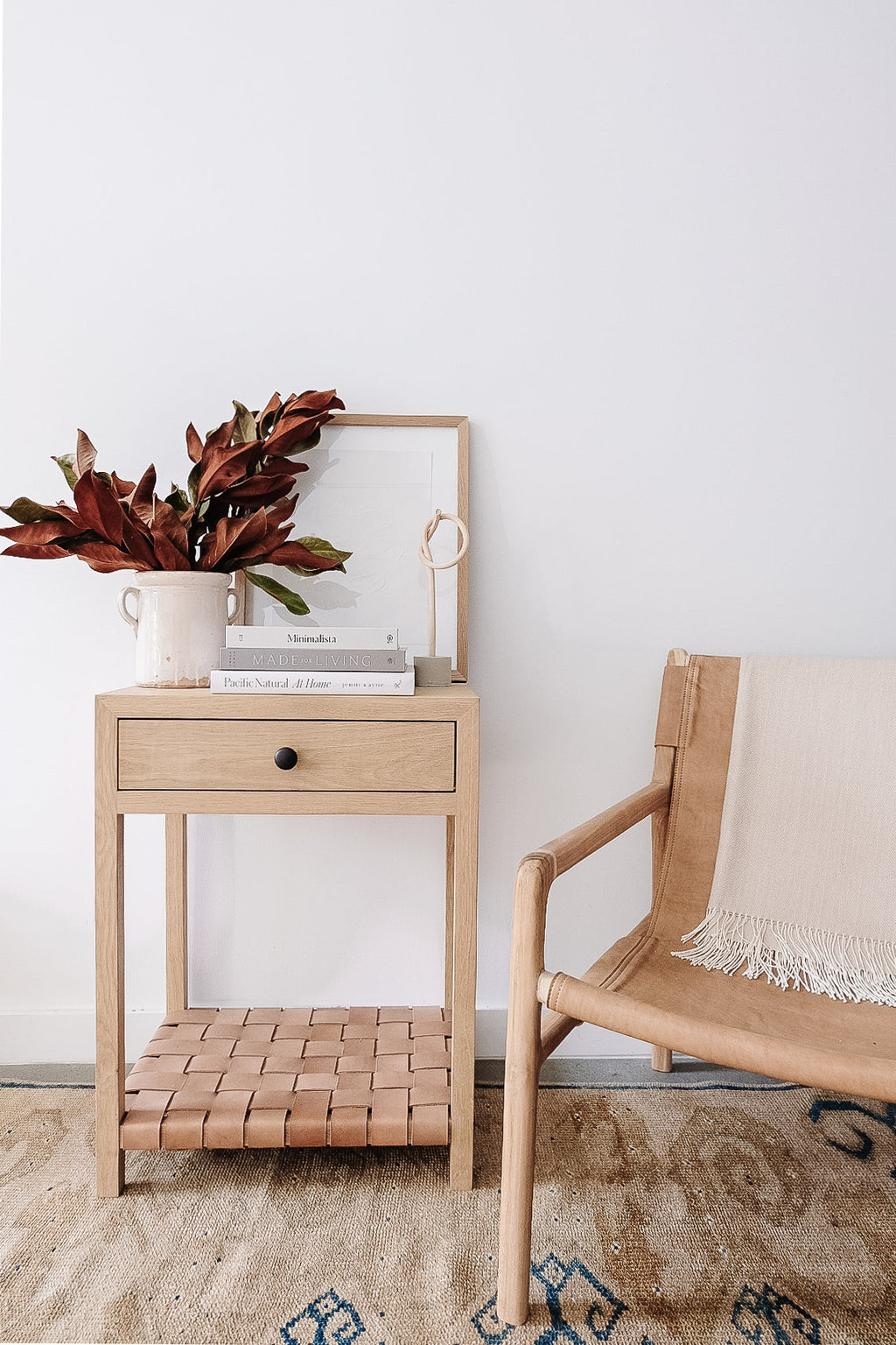 Minimalist wooden side table with dried leaves, books, and framed art next to a beige woven chair on a patterned rug