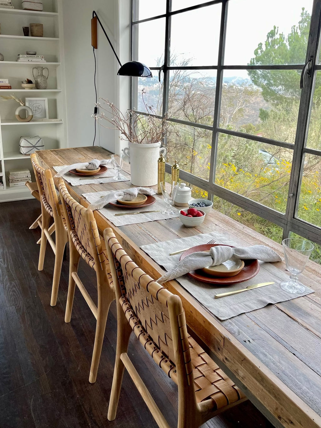 Rustic dining table set with natural linen placemats and wooden chairs near large windows
