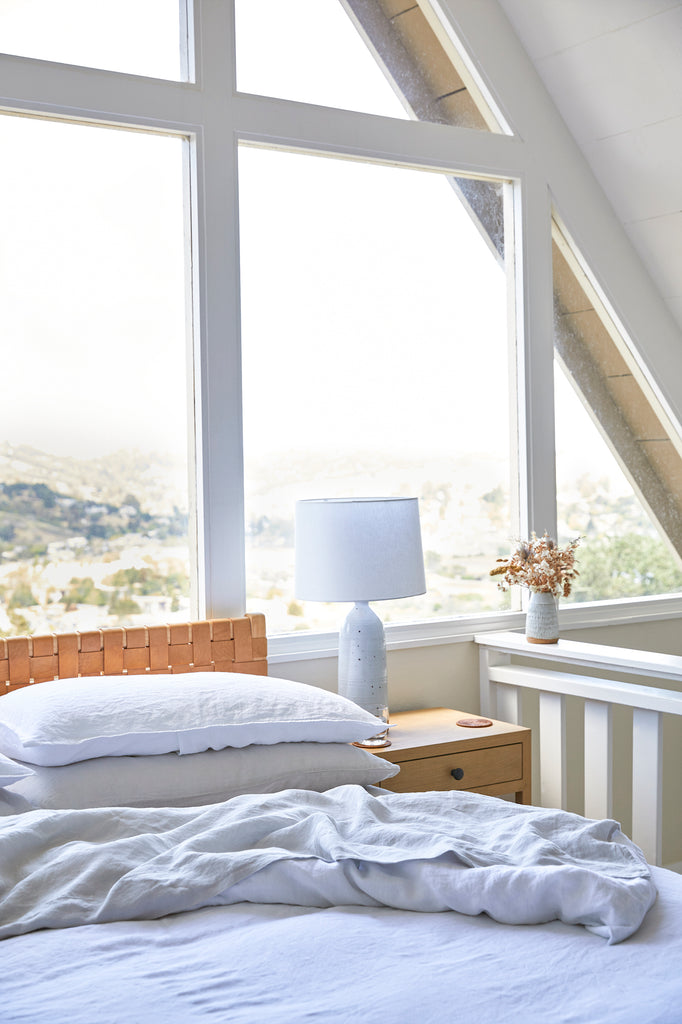 Styled Natural Linen Sheet Set with a Woven Leather Strap Headboard, an Oak and Leather Strap Side Table, a Tenganan Coaster, and a Hand Thrown Ceramic Lamp in an A-Frame home. - Saffron and Poe