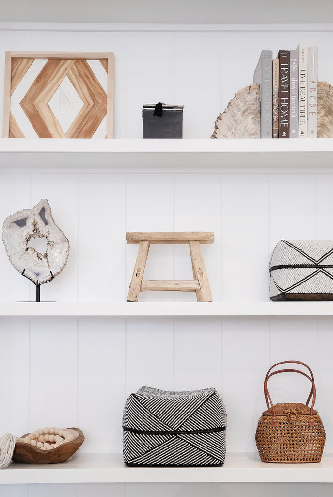 Styled petite rectangle stool on white shelf with hand beaded bali baskets, petrified wood bookends, tasseled bali beads, copper and teak bowl, ceramic canister, and Aleksandra Zee piece. - Saffron and Poe