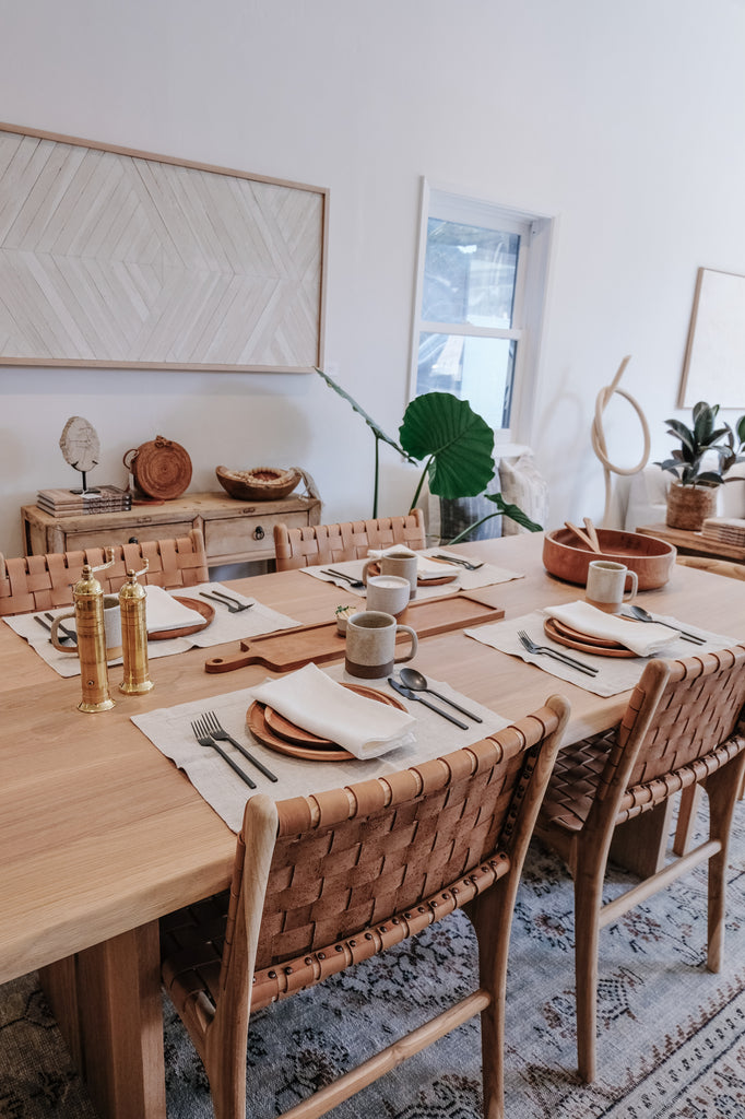 Styled Teak Plates with Linen Placemats, a Teak Salad Bowl, Uzumati Ceramic Mugs, Brass Grinders, and Woven Leather Strap Dining Chairs. - Saffron and Poe
