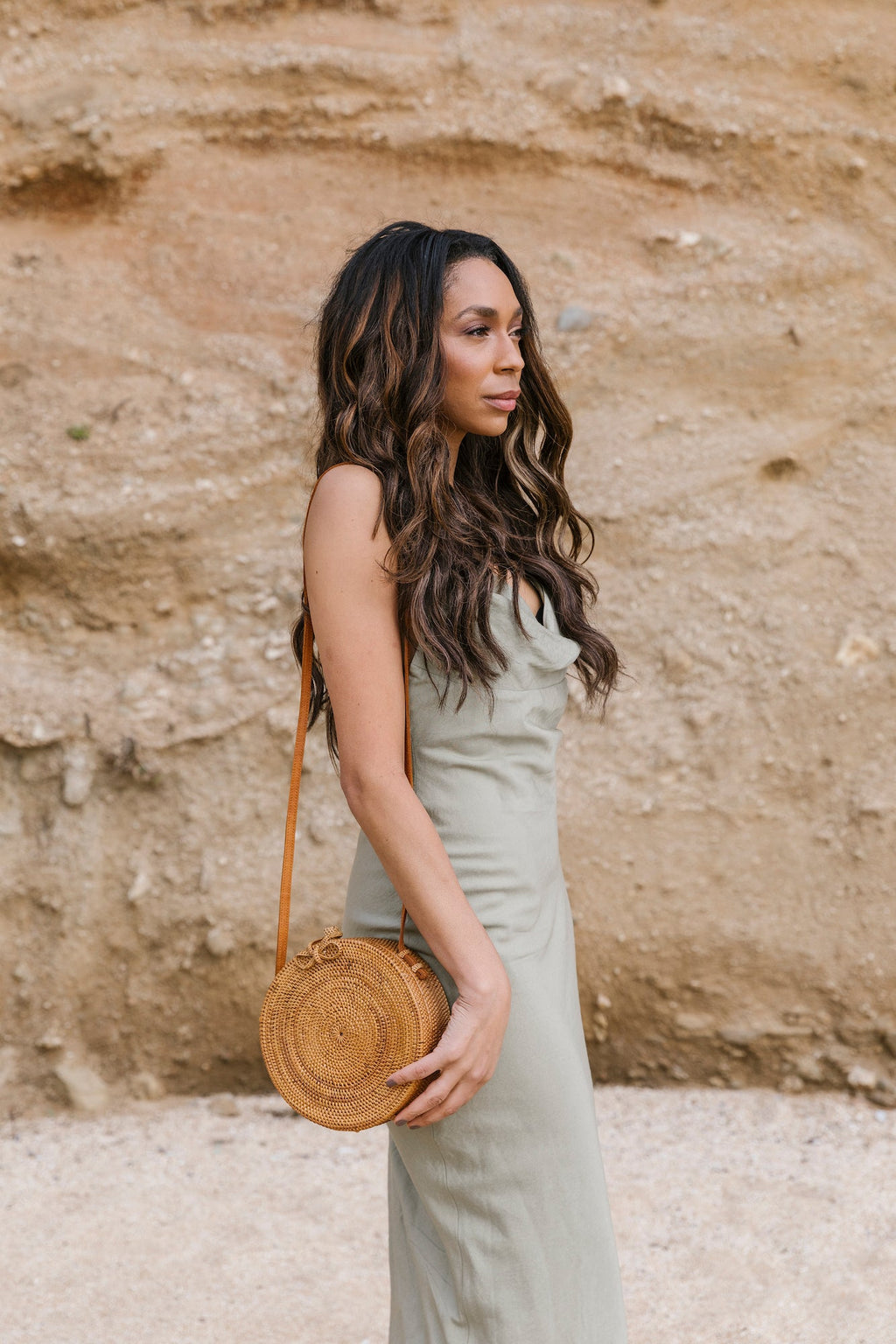 Woman wearing a light dress carrying a round Tenganan Basket Crossbody bag with woven bow closure against a natural rocky background
