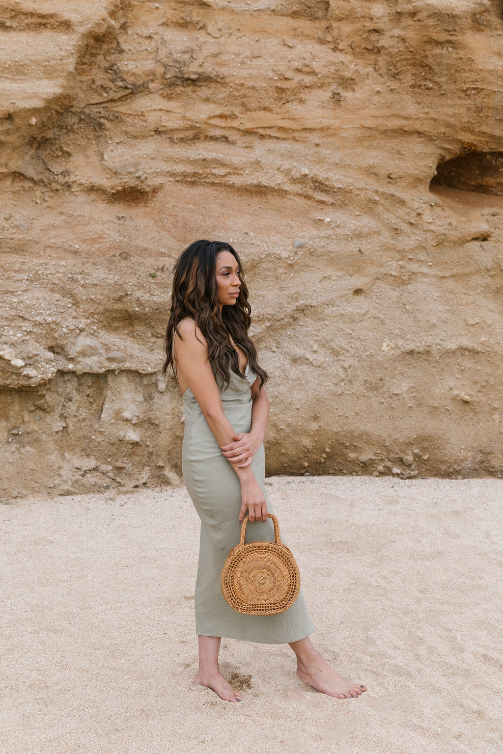 Woman holding Tenganan Basket Handbag made of natural Ata reed with intricate circular weave standing on sandy beach with rocky background