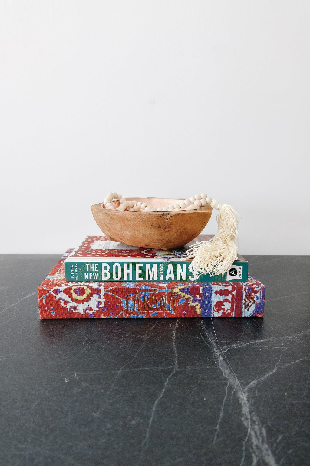 Stack of books including "The New Bohemians" on a dark surface with a wooden bowl and decorative tassel on top