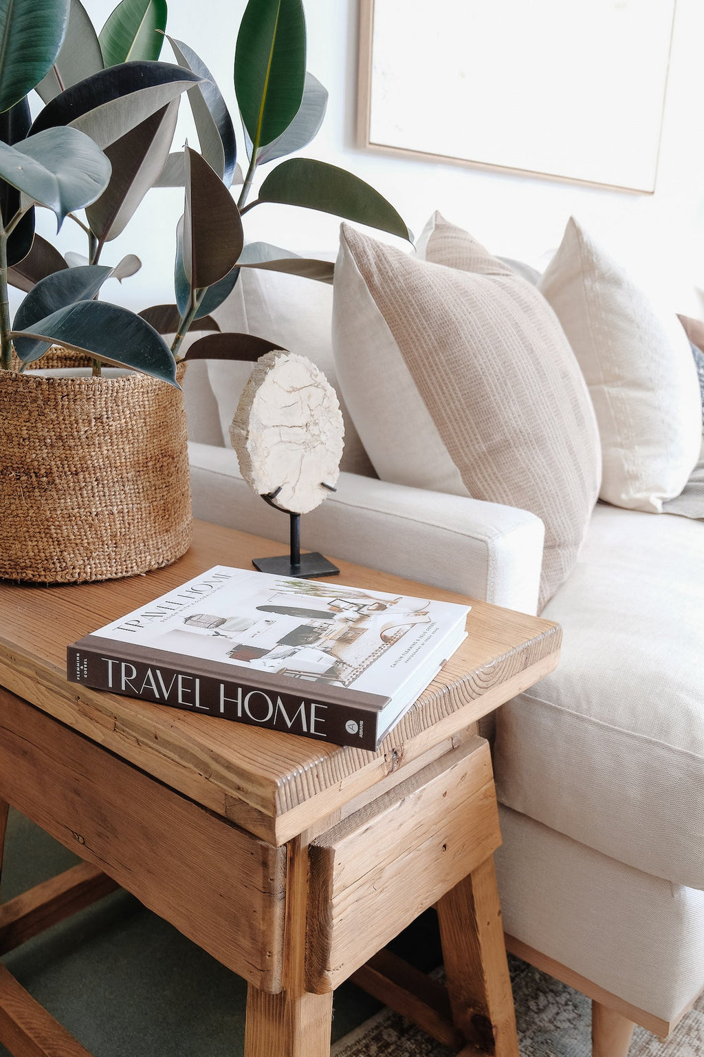 Travel Home book on wooden side table next to white couch with neutral pillows and large green potted plant.