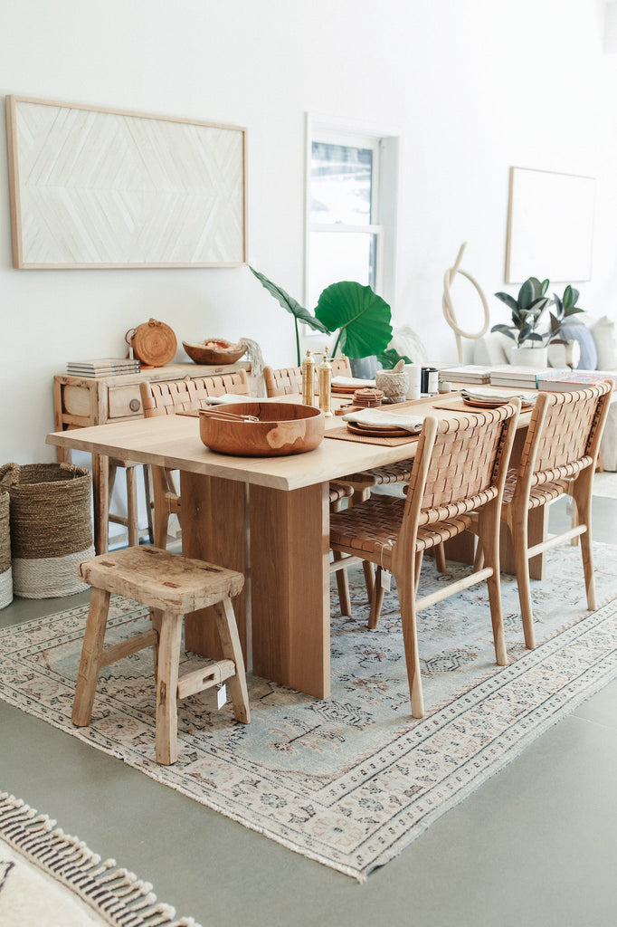 Styled vintage rectangular stool with white oak live edge table, vintage kilim rug, woven leather strap dining chairs in beige, green plant, Aleksandra Zee piece, round tenganan crossbody, teak wood serving bowl, Katie Gong Knot, and concrete floor with window. - Saffron and Poe