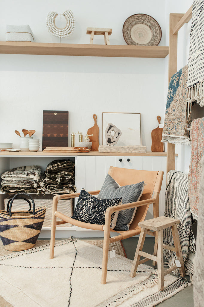 Styled vintage rectangular stool with leather sling back lounge chair, Jacob may cutting board, sculptural cutting boards, palm basket, shell necklace, petite rectangular stool, hand beaded bali basket, Moroccan Beni Ourain rugs, Indian bhujodi throws, oak shelving, black knobs, and white walls with antique hmong pillow and hemp textile pillow. - Saffron and Poe