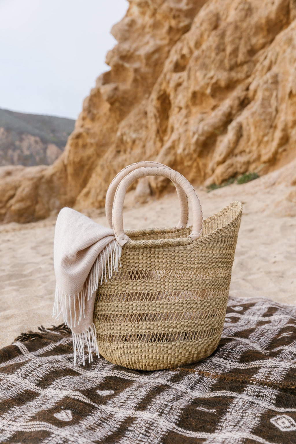 Woven Sisal Farmer's Market Tote with leather handles on a patterned blanket at the beach