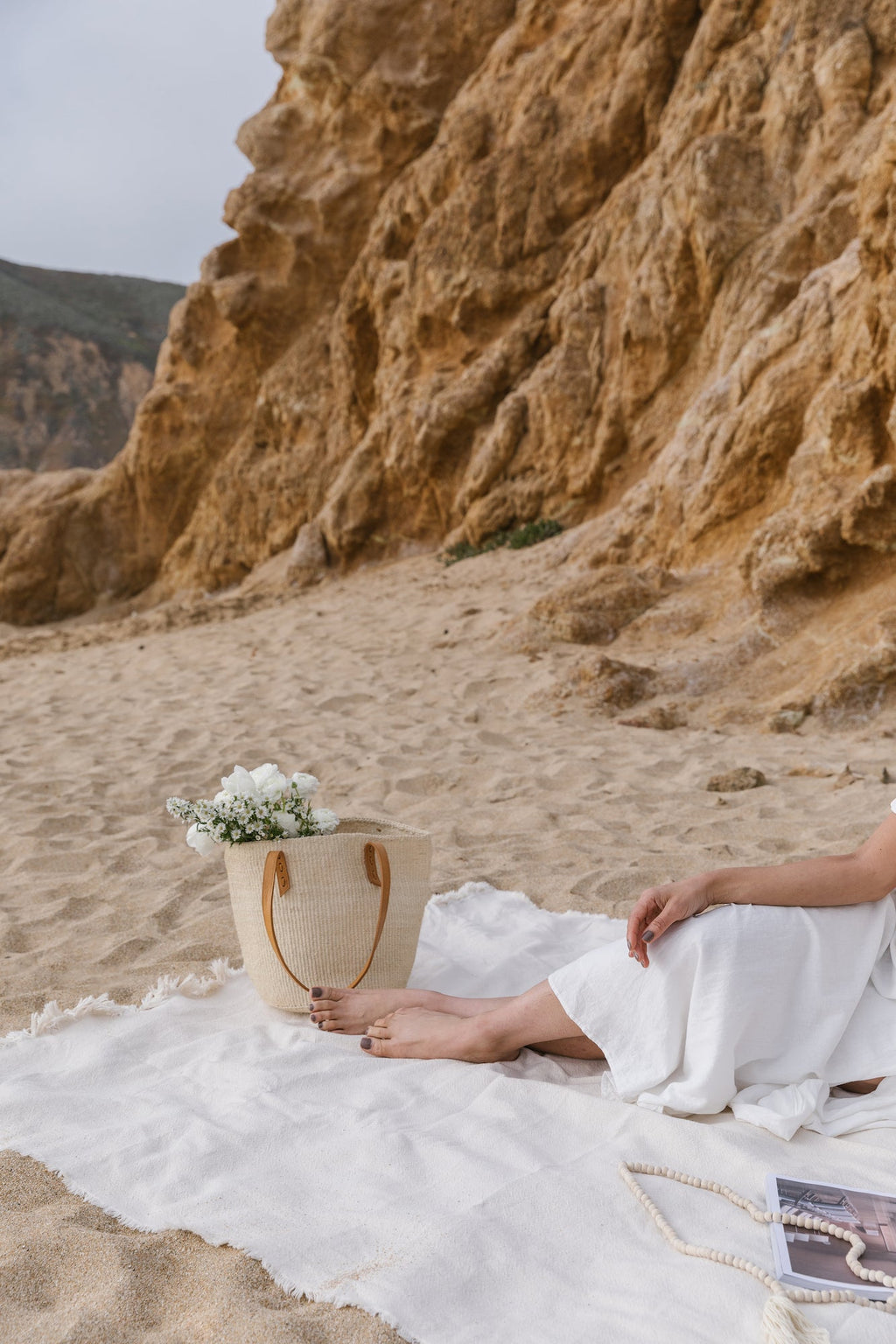 Woven sisal and leather tote bag on a sandy beach next to a person in a white dress sitting on a white blanket
