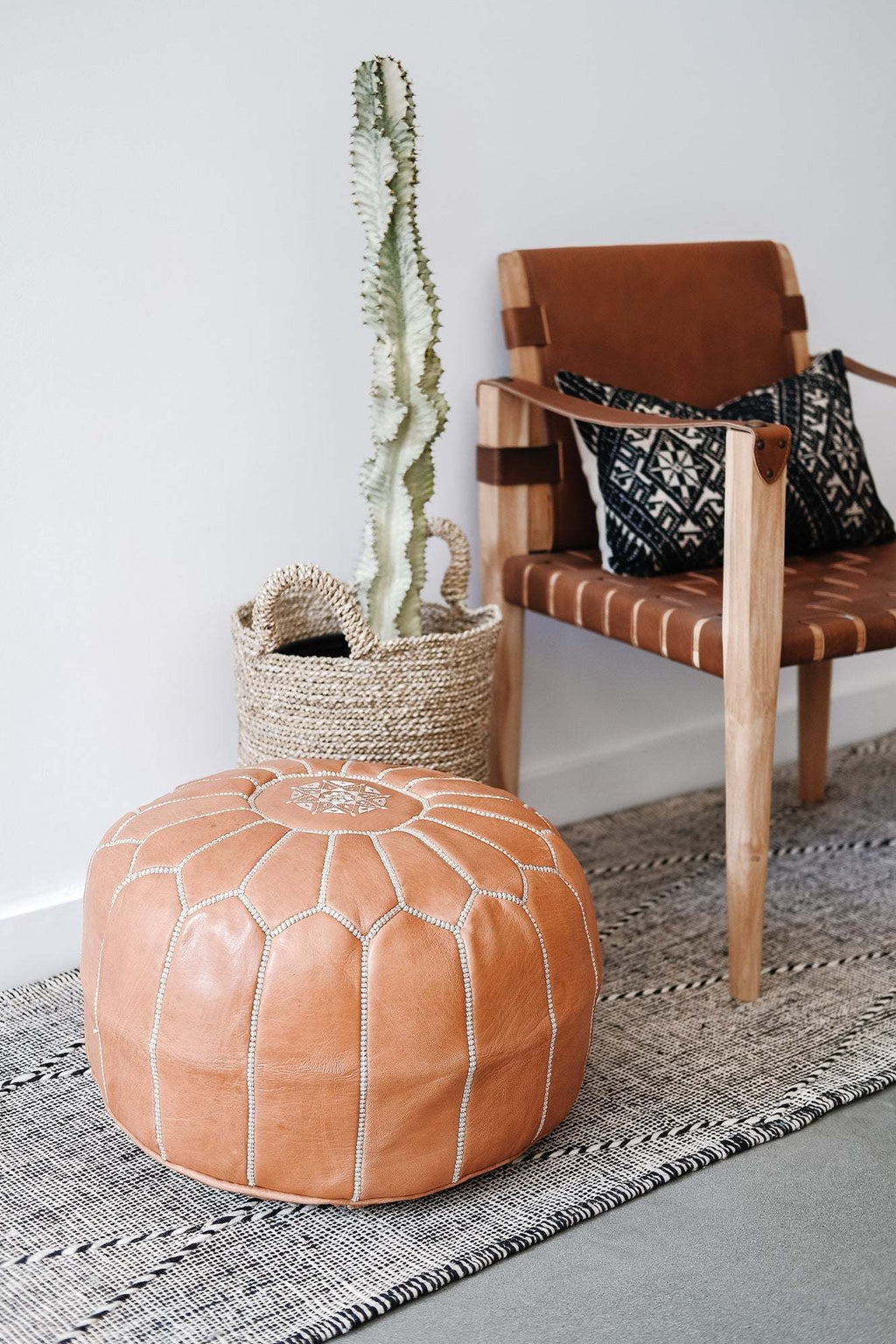 Embroidered Moroccan leather pouf in a modern living room setting with a wooden chair, patterned cushion, woven basket, and cactus plant.