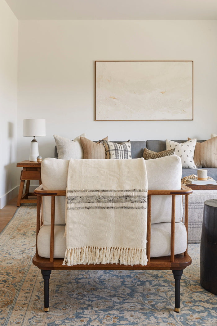 Styled view of our Handwoven Colombian Throw in grey draped over a decorative chair in a living room setting which includes our Ceramic Lamp, Vintage End Table, Bhujodi pillows, and Kaleen Cameron art work. - Saffron and Poe.