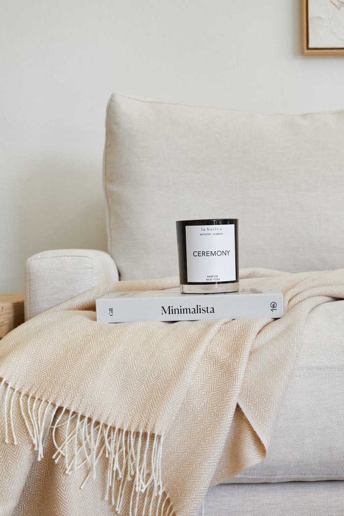 Styled view of Winter Bundle which includes Beige Baby Alpaca Throw, La Botica Ceremony Candle, and Minimalista book on a cream sofa against a white background. - Saffron and Poe