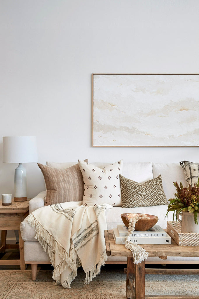 Styled close up view of our Handwoven Colombian Throw in grey draped over sofa with decorative pillows, Ceramic Lamp, Vintage Coffee Table, Vintage Side Table, Aleksandra Zee art work, and Turkish Rug. - Saffron and Poe