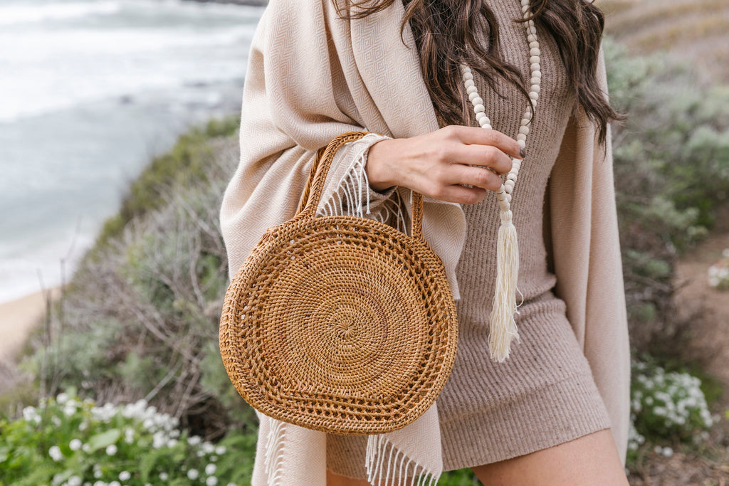 Woman wearing beige baby alpaca throw shawl holding a round woven handbag near the beach